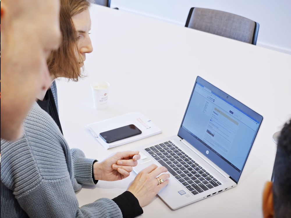 Twee collega's werken samen aan een laptop op een witte tafel; de vrouw gebruikt het touchpad en er ligt een notitieblok en een smartphone naast.