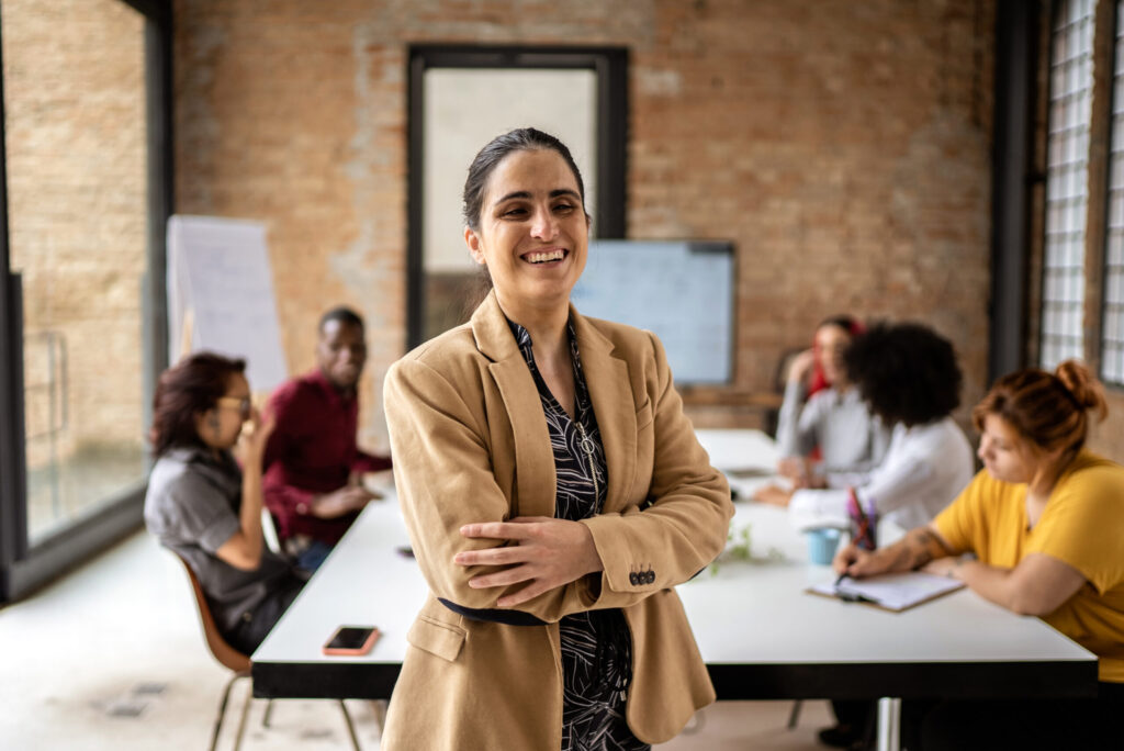 Vrolijke, professionele vrouw met een visuele beperking en gekruiste armen, staand voor een team dat vergadert aan een grote tafel.