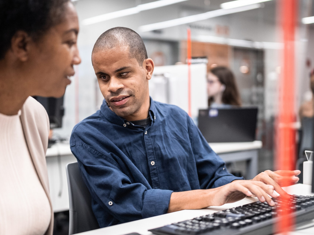Een man met een visuele beperking typt op een toetsenbord terwijl hij praat met een collega die naast hem zit.