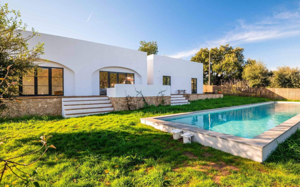 Een moderne, witte vakantiewoning met een strak zwembad in een groene tuin onder een blauwe lucht.