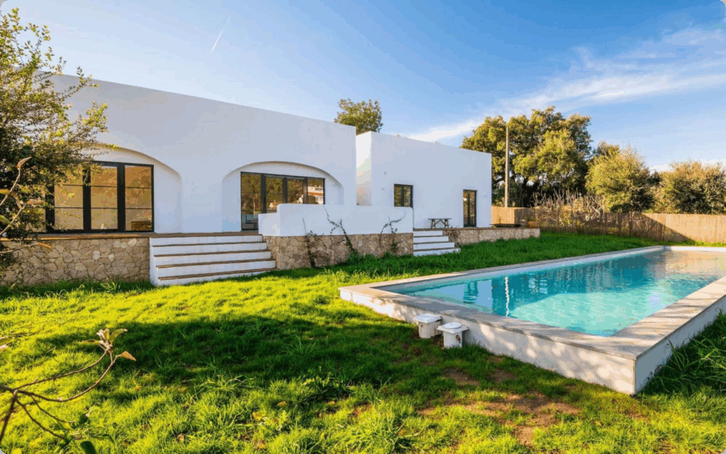 Een moderne, witte vakantiewoning met een strak zwembad in een groene tuin onder een blauwe lucht.