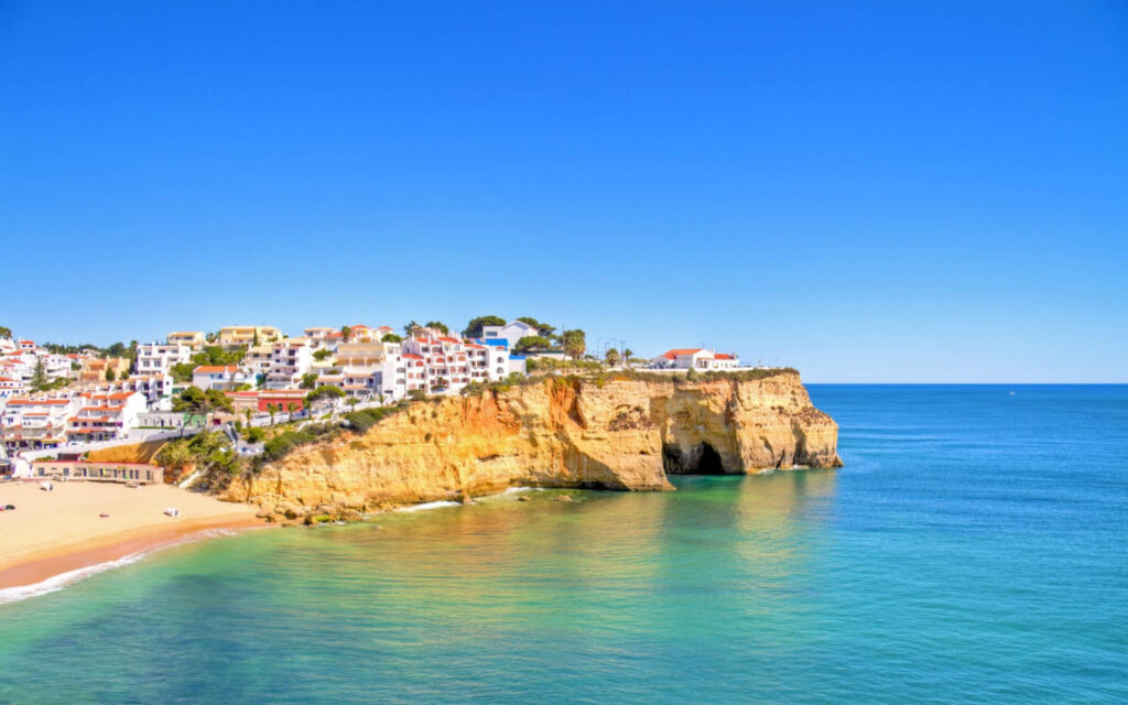 Een kustlijn met witte huizen op een rotswand boven een goudgeel strand en een helderblauwe zee.