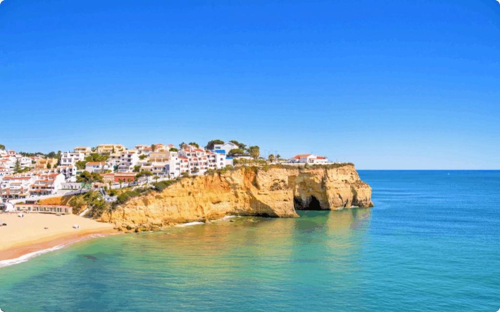 Een kustlijn met witte huizen op een rotswand boven een goudgeel strand en een helderblauwe zee.