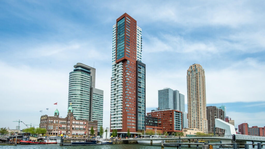 Een stadsgezicht vanaf het water met diverse moderne woontorens en het historische Hotel New York aan de kade.