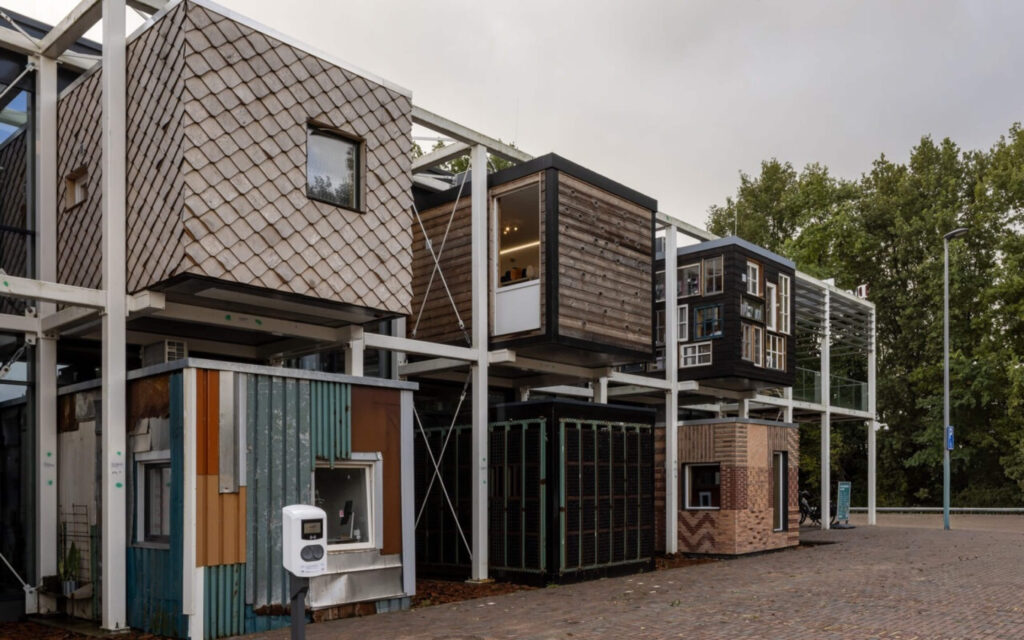 Een witte staalconstructie met diverse modulaire units, elk met een andere gevel zoals hout, zwarte kozijnen of gekleurde golfplaten.