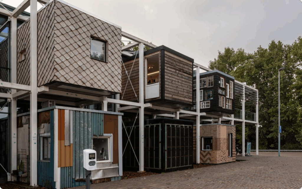 Een witte staalconstructie met diverse modulaire units, elk met een andere gevel zoals hout, zwarte kozijnen of gekleurde golfplaten.