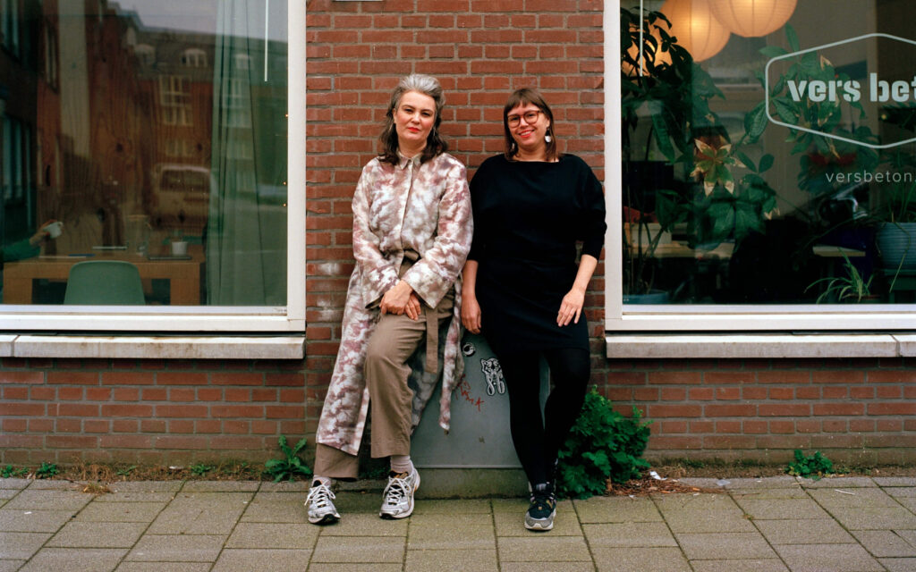 Eeva Liukku en uitgever Hilde Westerink staan glimlachend buiten tegen een bakstenen muur tussen twee ramen van een kantoor; op het rechterraam staat het logo van 'Vers Beton'.