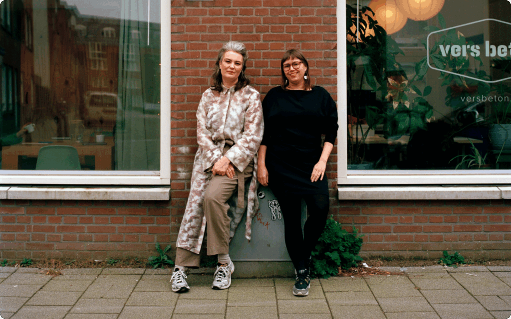 Eeva Liukku en uitgever Hilde Westerink staan glimlachend buiten tegen een bakstenen muur tussen twee ramen van een kantoor; op het rechterraam staat het logo van 'Vers Beton'.