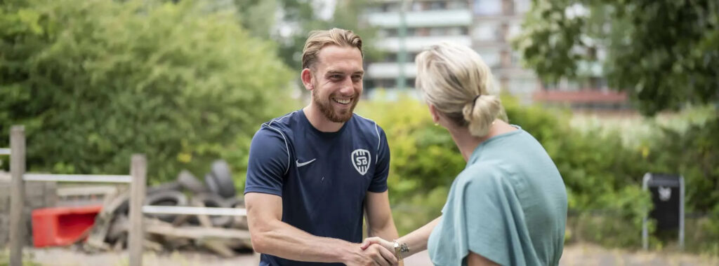 Een lachende man in een donkerblauw SBG-shirt schudt de hand van een blonde vrouw met het haar in een knot, waarschijnlijk na een succesvolle samenwerking.