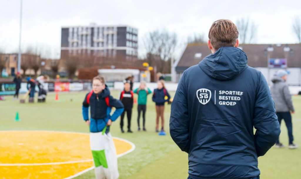 Een mannelijke sportcoach in een donkerblauwe jas met het logo 'Sportief Besteed Groep' op de rug kijkt toe hoe kinderen zakdoeklopen of sporten op een grasveld.