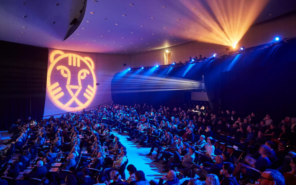 Een grote bioscoopzaal gevuld met publiek, verlicht door blauwe sfeerverlichting en een projectie van de IFFR-tijger op de zijwand.