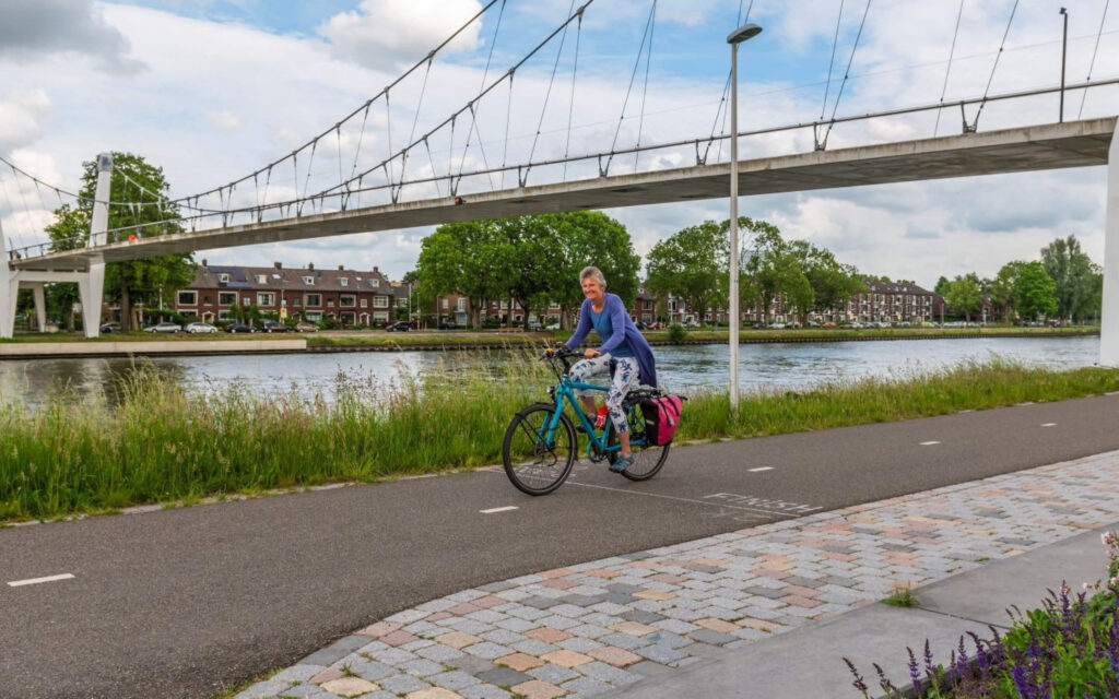Een vrouw fietst op een blauwe toerfiets met roze fietstassen over een geasfalteerd fietspad langs een kanaal, met een grote wit-grijze hangbrug op de achtergrond.