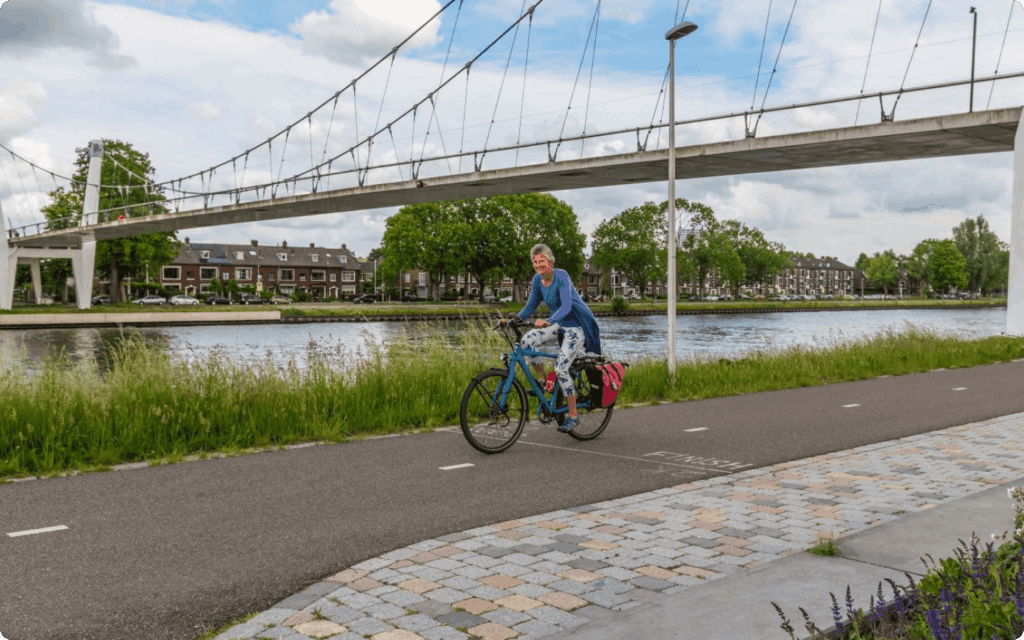 Een vrouw fietst op een blauwe toerfiets met roze fietstassen over een geasfalteerd fietspad langs een kanaal, met een grote wit-grijze hangbrug op de achtergrond.