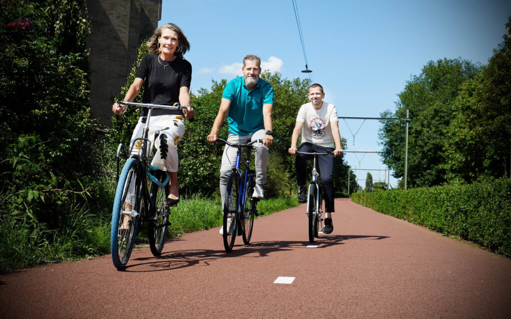 Drie volwassenen fietsen glimlachend naast elkaar op een breed rood fietspad in een groene, bosrijke omgeving onder een strakblauwe lucht.