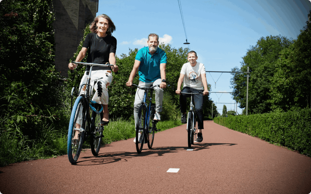 Drie volwassenen fietsen glimlachend naast elkaar op een breed rood fietspad in een groene, bosrijke omgeving onder een strakblauwe lucht.