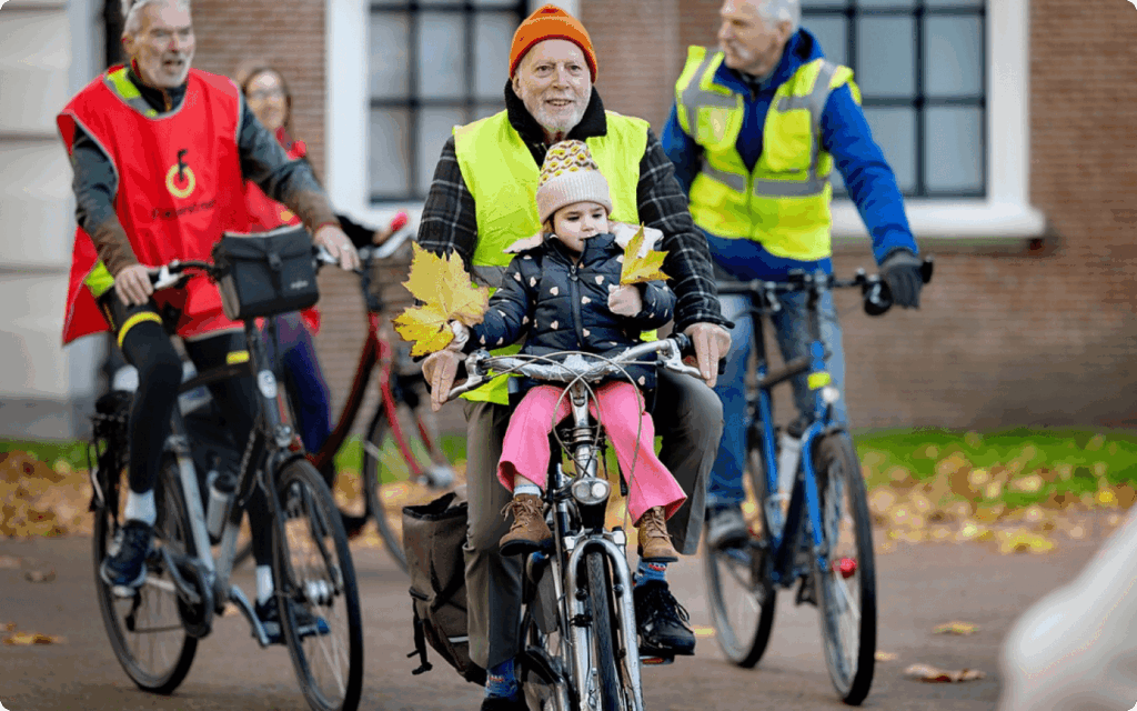 Een man fietst met een jong kind in een voorzitje tijdens een herfstige fietstocht; de man draagt een reflecterend veiligheidshesje en het kind houdt herfstbladeren vast.