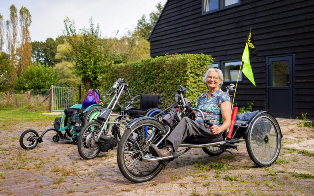 Een lachende vrouw zit op een aangepaste driewielfiets voor een zwart houten gebouw, met naast haar diverse andere modellen aangepaste fietsen en rolstoelen.