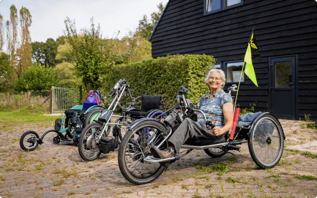 Een lachende vrouw zit op een aangepaste driewielfiets voor een zwart houten gebouw, met naast haar diverse andere modellen aangepaste fietsen en rolstoelen.
