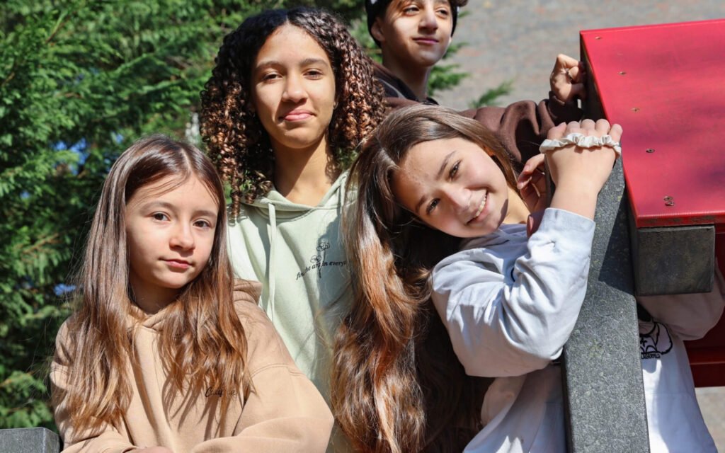 Groepsportret van vier lachende jongeren van verschillende leeftijden die buiten bij een houten object poseren in het zonlicht.
