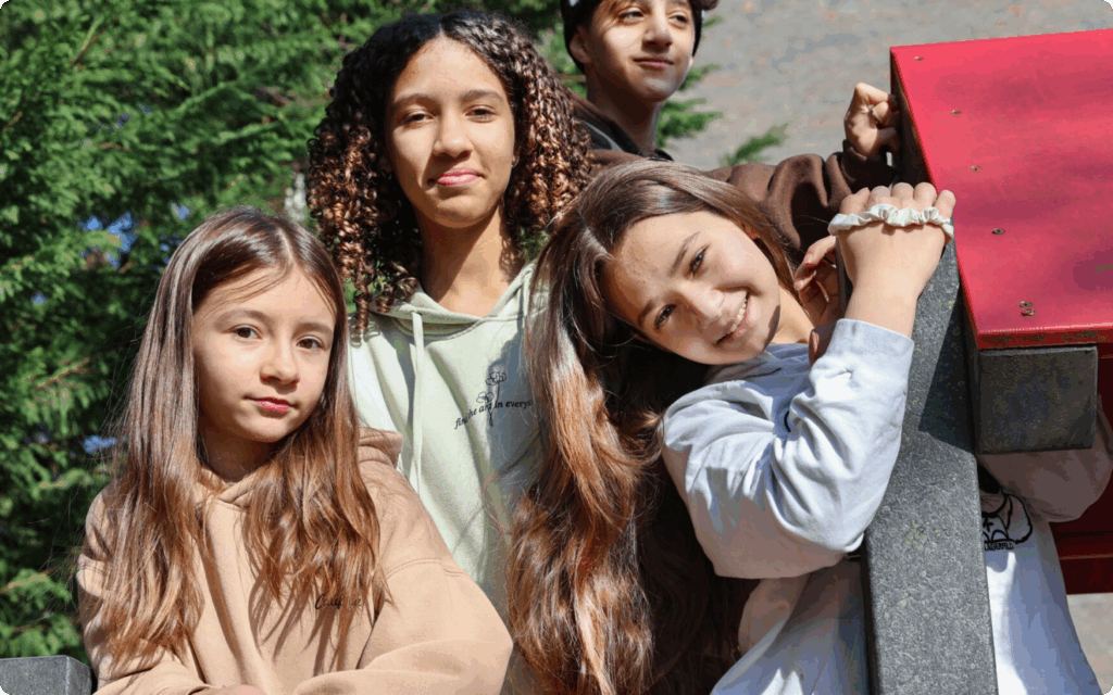 Groepsportret van vier lachende jongeren van verschillende leeftijden die buiten bij een houten object poseren in het zonlicht.
