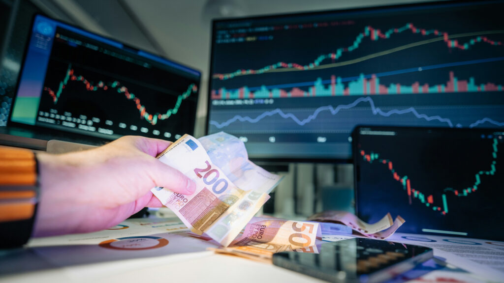 Een hand met een biljet van 200 euro voor computerschermen met beursgrafieken.