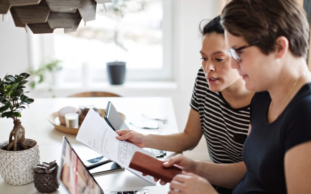Twee vrouwen zitten samen aan een tafel en bestuderen aandachtig een officieel document bij een laptop.