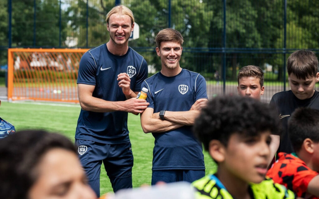 Twee glimlachende voetbaltrainers in donkerblauwe sportkleding met een wit logo van SBG staan op een groen voetbalveld tijdens een training voor een groep kinderen.