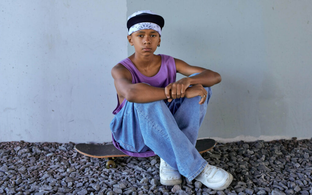 Een jongen met een bandana en pet zit met gevouwen handen op een skateboard voor een grijze muur.