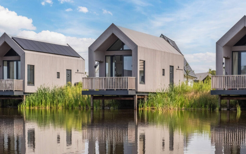 Een rij moderne, houten recreatiewoningen op palen boven het water, voorzien van zonnepanelen op de daken.