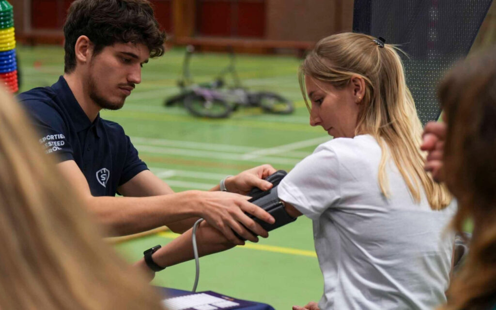 Een mannelijke sportprofessional in een donkerblauwe SBG-polo meet de bloeddruk van een jonge blonde vrouw tijdens een vitaliteitsdag of gezondheidscheck.