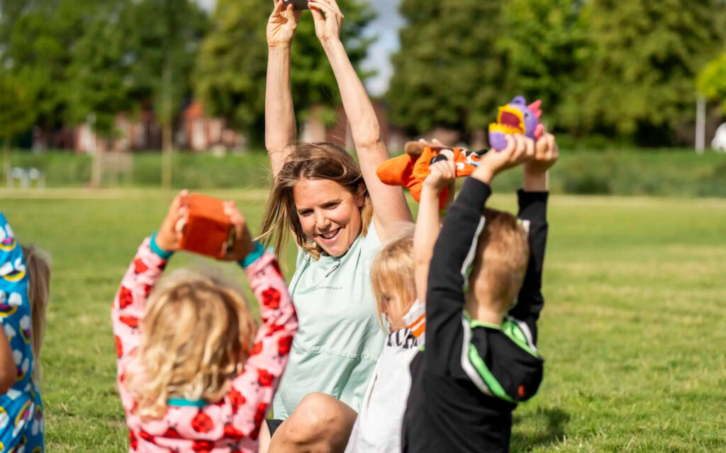 Vrolijke buurtsportcoach begeleidt jonge kinderen met speelse sportactiviteiten op een grasveld.
