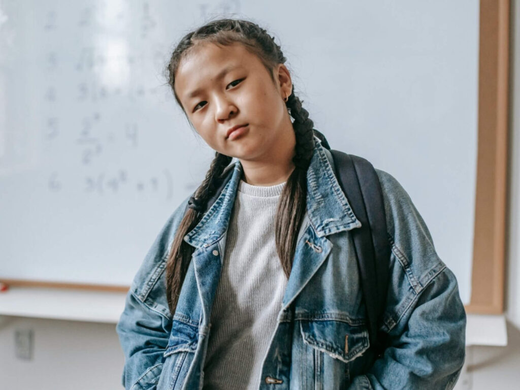 Portret van een meisje met twee vlechten en een spijkerjasje die met een serieuze blik voor een wit schoolbord staat.