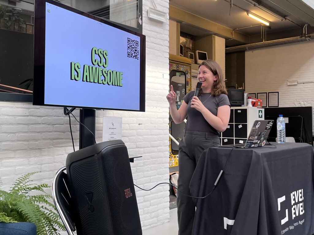 Een vrouw met microfoon wijst naar een beeldscherm waar de woorden "CSS is Awesome" te lezen zijn.