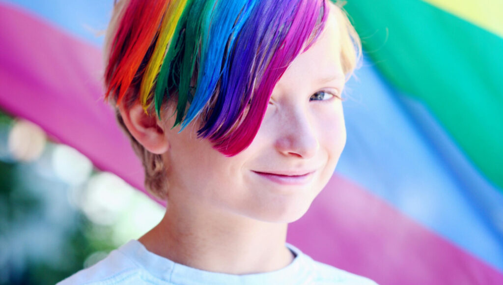 Een close-up portret van een kind dat glimlacht met opvallend regenboogkleurig geverfd haar, tegen de achtergrond van een wapperende regenboogvlag.