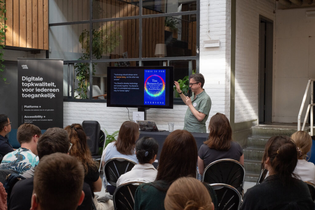 Een man staat voor publiek en geeft een presentatie. Hij wijst met een hand naar een scherm waar het boek "This is for Everyone" van Tim Berners-Lee te zien is.