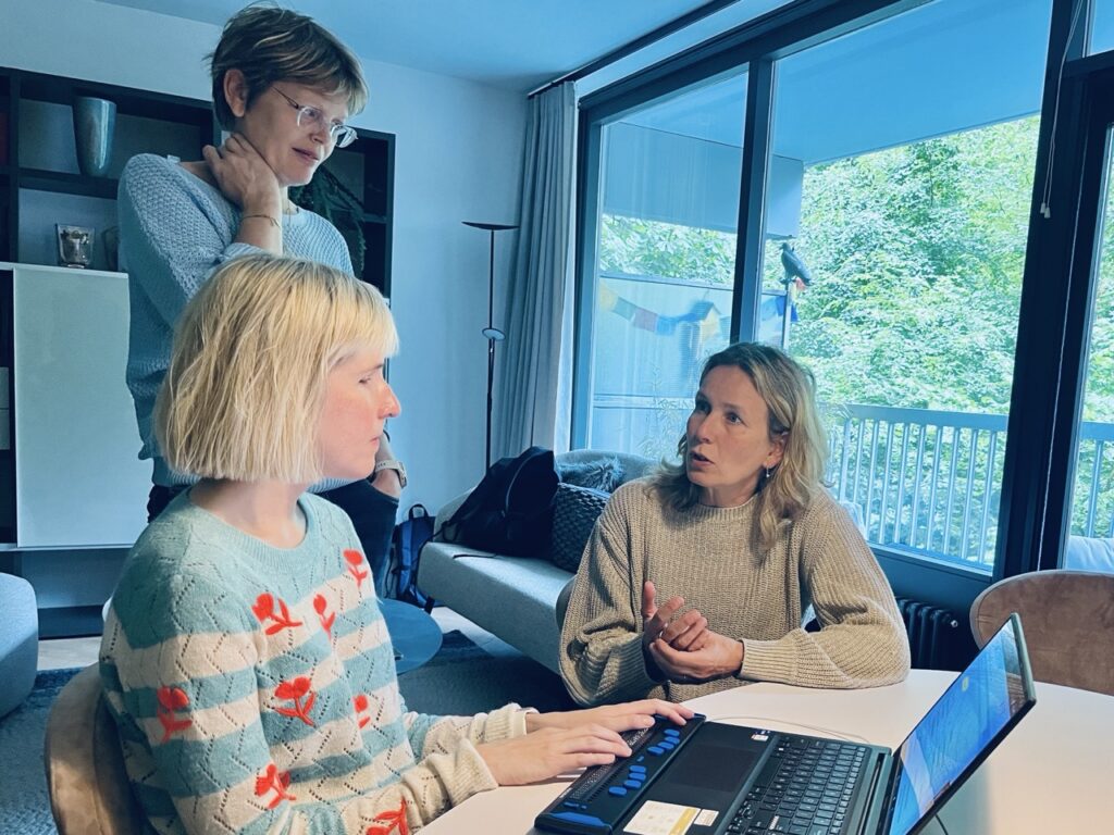 Drie vrouwen in een woonkamer met een groot raam, buiten zijn bomen en een stuk balkon te zien. Twee vrouwen zitten aan tafel en praten met elkaar. Een heeft een opengeklapte laptop voor zich en leest met haar vingers de braille-leesregel. Een vrouw staat er achter en luistert.