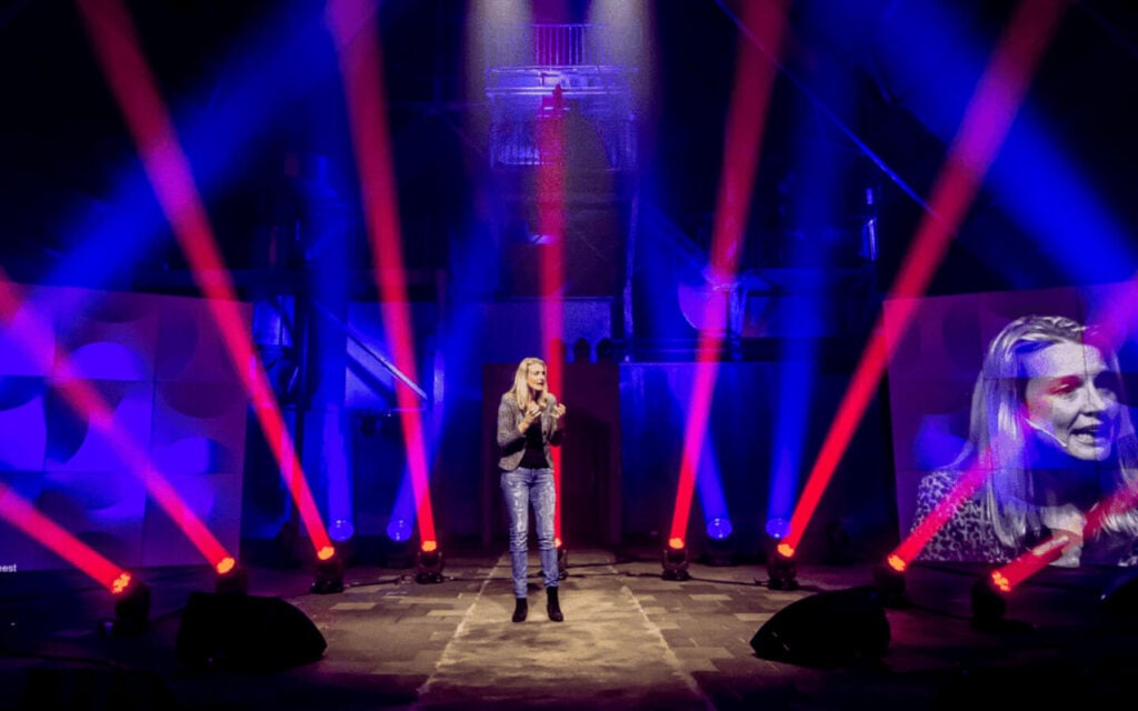 Spreker op een podium verlicht door felle rode en blauwe schijnwerpers.