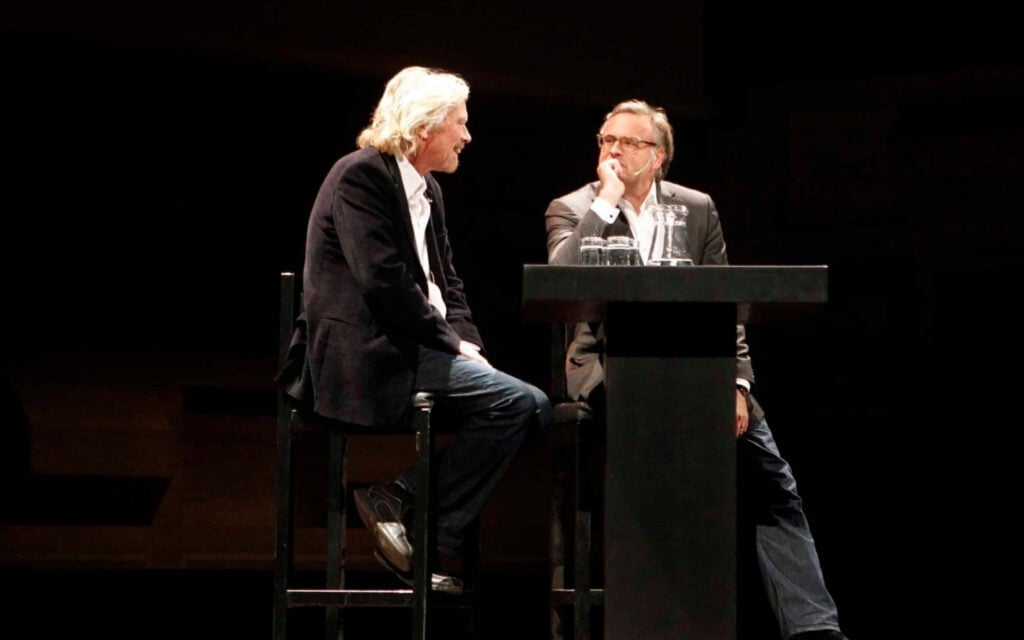 Twee sprekers op hoge stoelen in gesprek op een donker podium.