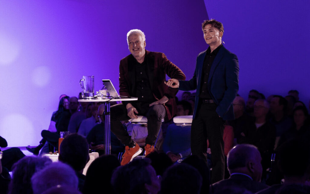 Twee sprekers op een paars belicht podium voor een zaal met publiek.