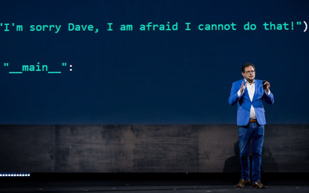Spreker op een podium voor een groot scherm met geprojecteerde programmeercode.
