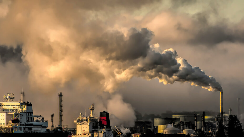 Dichte rook- en stoomwolken stijgen op uit een fabriekspijp van een industrieel complex bij een haven met schepen.