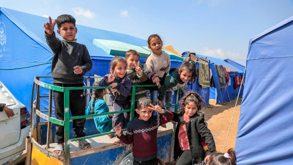 Een groep lachende kinderen staat op een karretje tussen opvangtenten en maakt het peace-teken naar de camera.