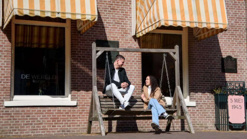 Twee jongvolwassenen zitten pratend op een houten schommelbank voor hotel-restaurant Hotel De Wereld.