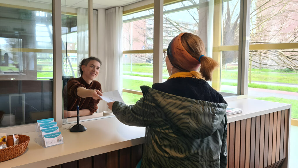 Een bezoeker overhandigt een document aan een medewerker achter een balie met een glazen wand.